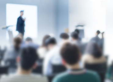 A blurred styled photo of a conference room filled with people with a presenter in the front of the room.