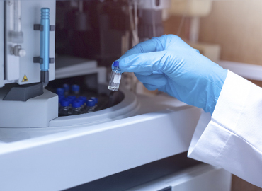 A gloved hand grabs a vial from a machine in a laboratory.