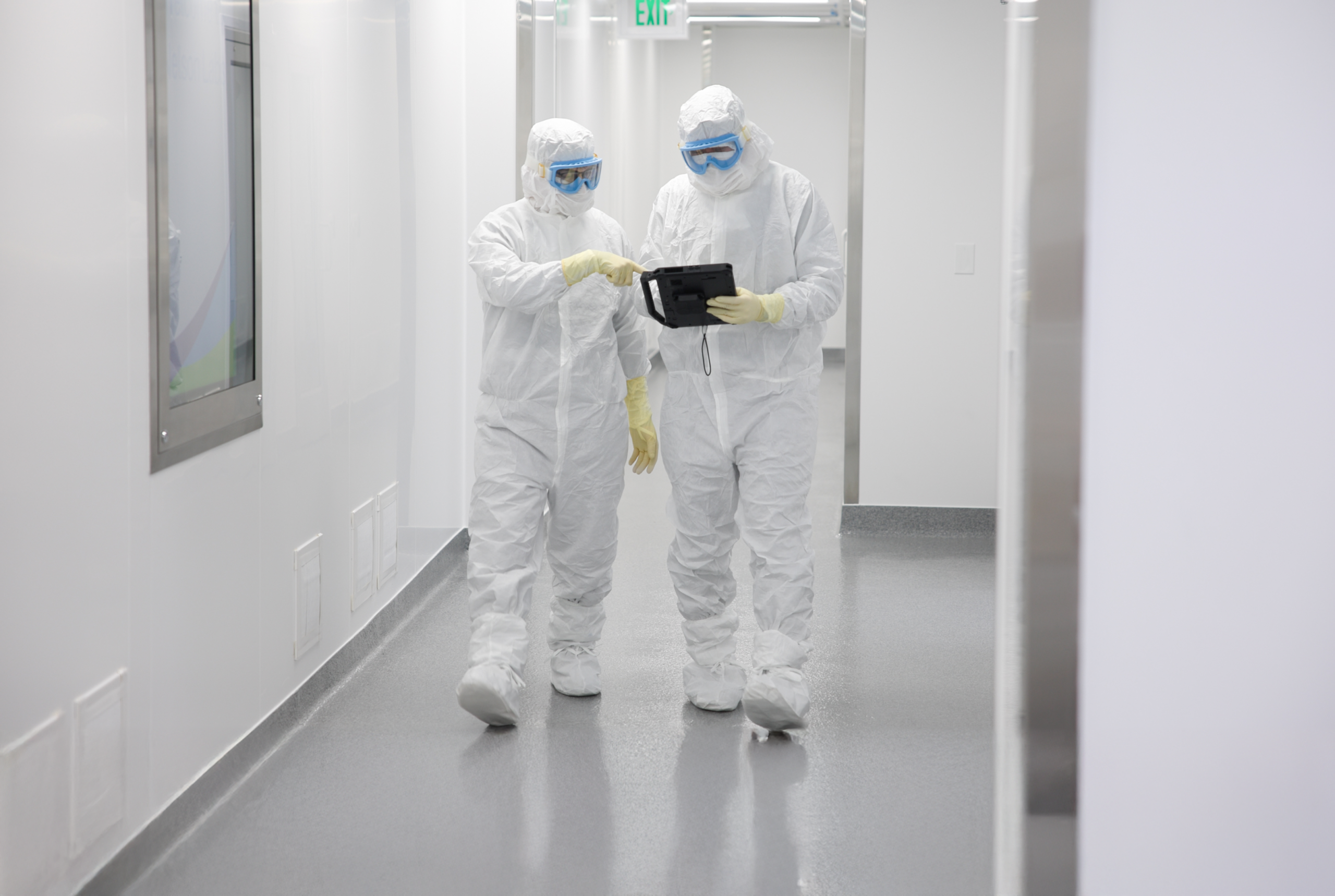 Two laboratory workers in full protective hazmat suits looking at a tablet in a cleanroom hallway