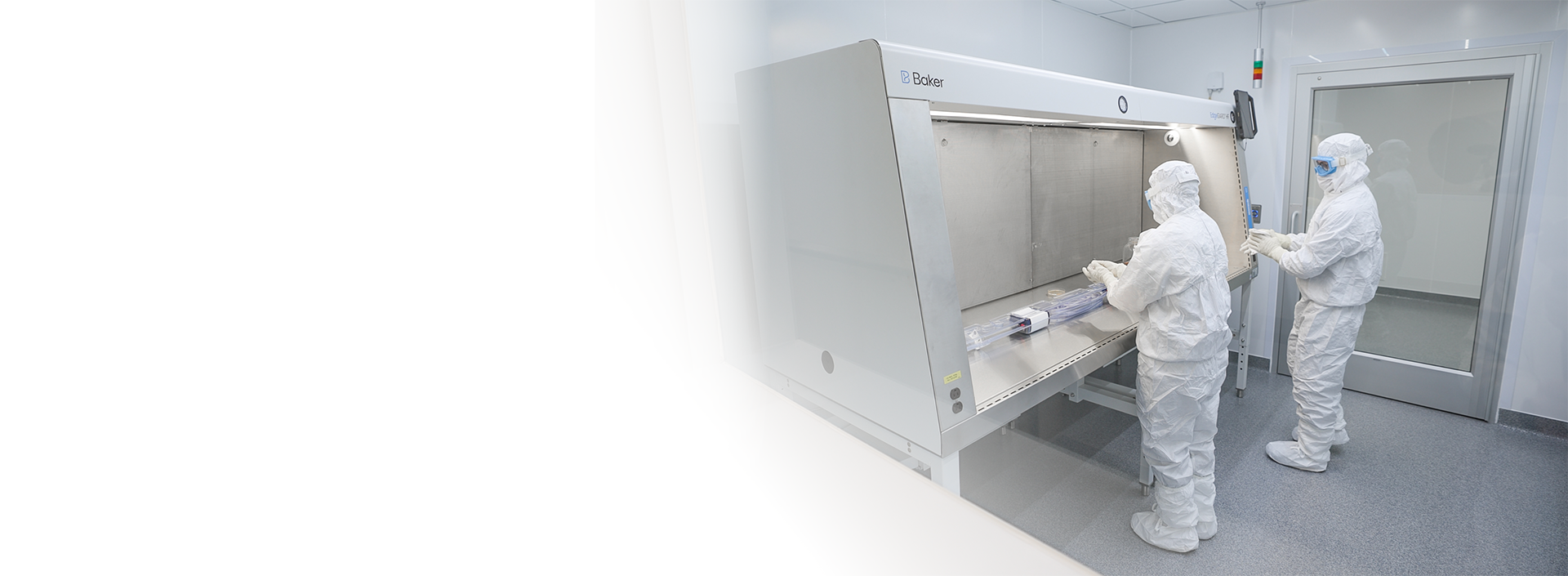 Panoramic banner graphic showing two laboratory technicians in white protective cleanroom suits working at a large Baker biosafety hood, with the left side of the image fading completely into solid white.