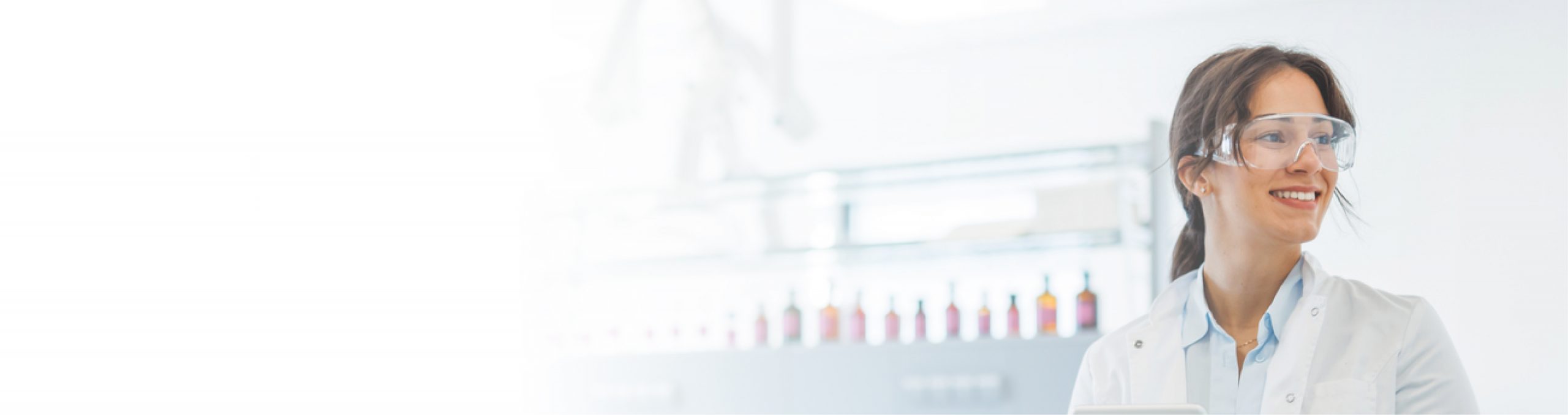 A panoramic, high-key banner image featuring a smiling female scientist positioned on the right side, wearing a white lab coat and clear safety glasses while looking off-camera. The background shows a bright, out-of-focus laboratory environment with a row of small bottles on a shelf, seamlessly fading into a solid white space on the left side of the frame.
